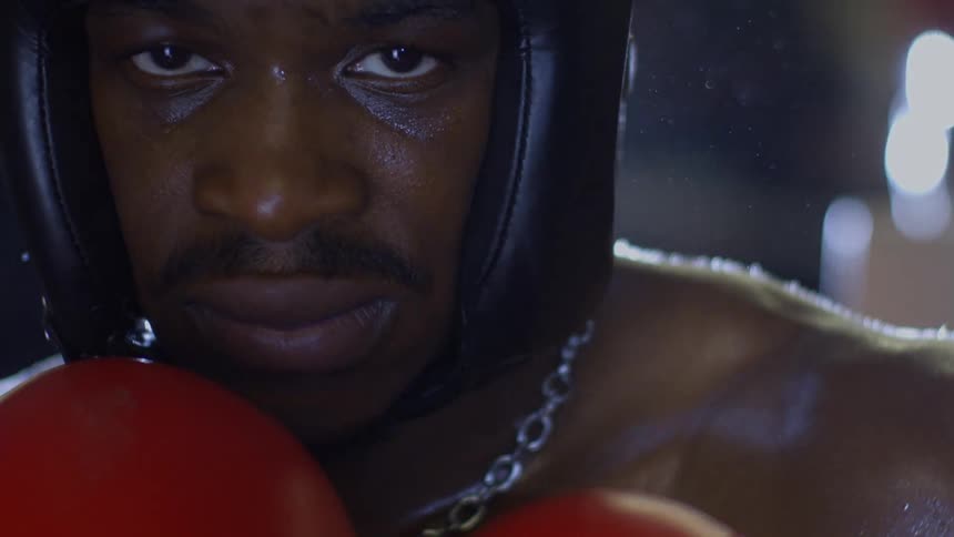 An intense and sweaty boxer in a head guard and red gloves gives a focused stare in a dramatic slow-motion close-up