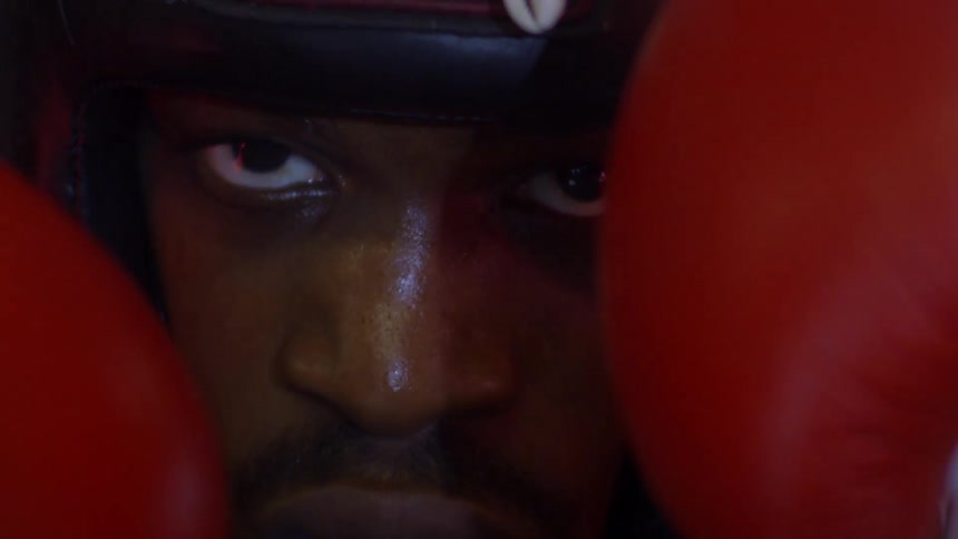 An intense and determined boxer in protective headgear stares directly at the camera in an extreme close-up