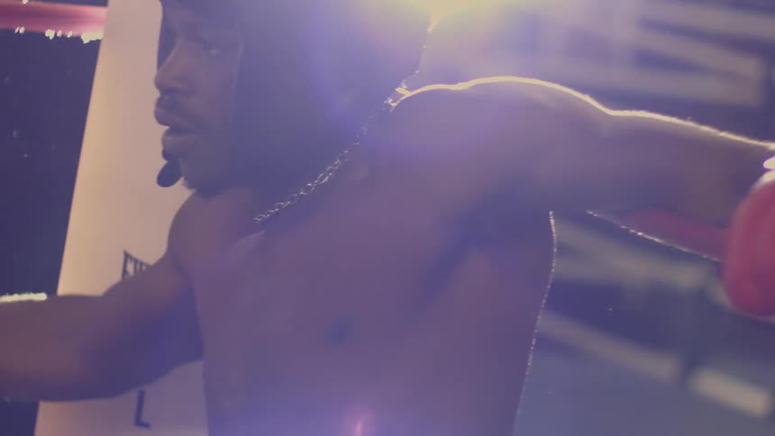 A determined shirtless boxer with headgear leans on the ropes, catching his breath in the corner of a brightly lit ring in slow motion