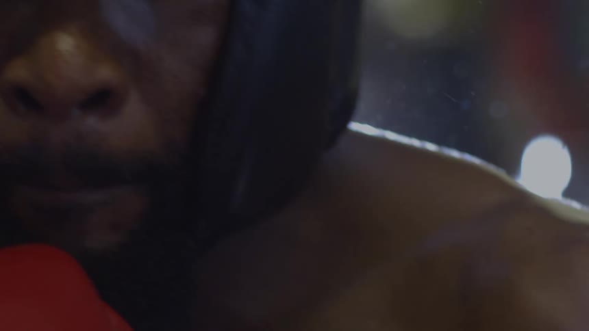 Intense slow-motion close-up of a determined boxer's face with headgear, staring forward in a dramatically lit gym