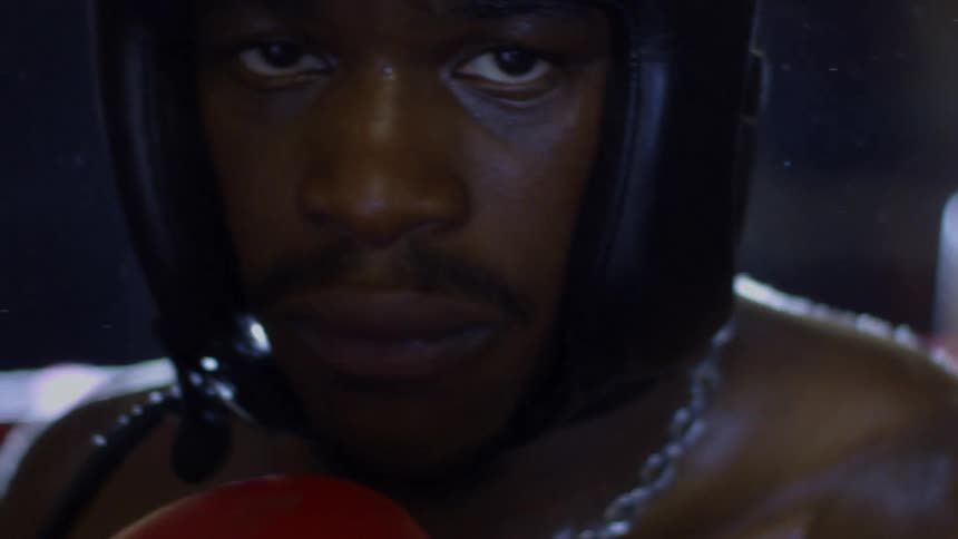 A determined male boxer in protective headgear stares intensely at the camera in a dramatic slow-motion close-up