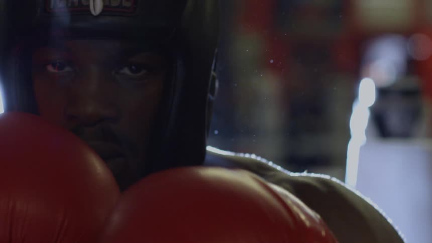 Dramatic slow-motion close-up of a determined boxer in full gear, staring at the camera before his red glove moves to cover the lens
