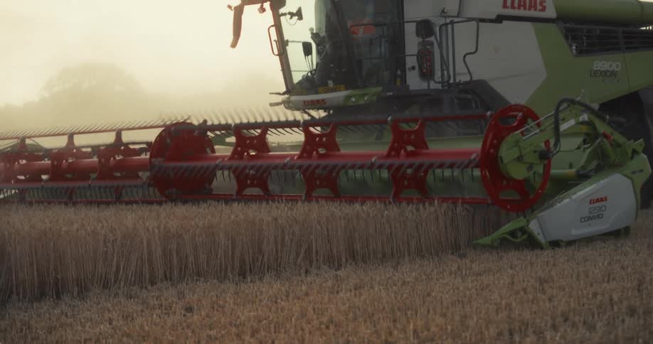 A combine harvester's spinning reel cuts through a golden wheat field in slow motion during a hazy sunset