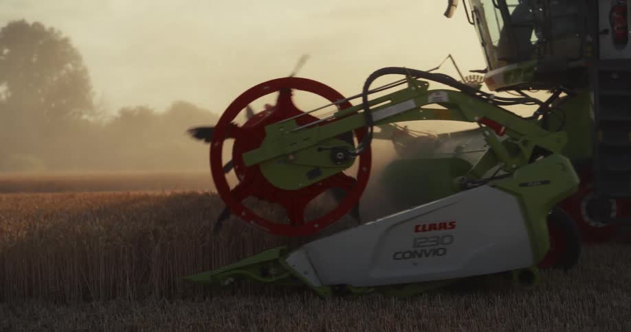 Close-up slow-motion shot of a combine harvester cutting through a golden wheat field during a dusty sunset harvest