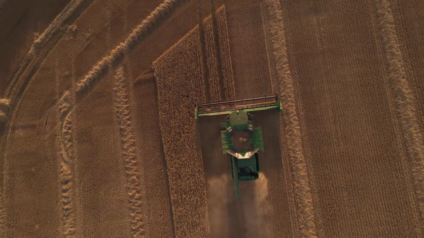 A top-down aerial view shows a combine harvester working in a golden wheat field, leaving patterns as it reaps the crops