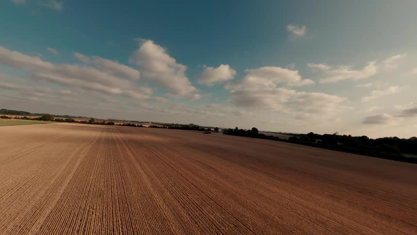 An expansive aerial shot drifts over a vast, golden-brown farmland under a partly cloudy sky