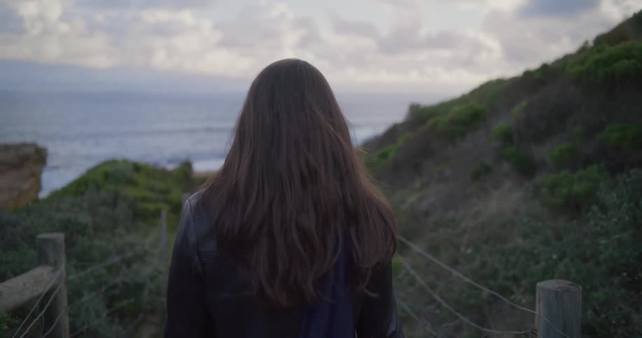 A solitary woman with long, flowing brown hair walks down a tranquil coastal path toward the ocean at dusk