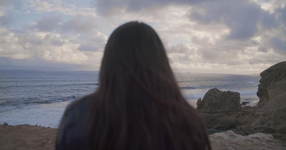 Over-the-shoulder shot of a woman with long brown hair watching the ocean waves from a rocky cliff at dusk