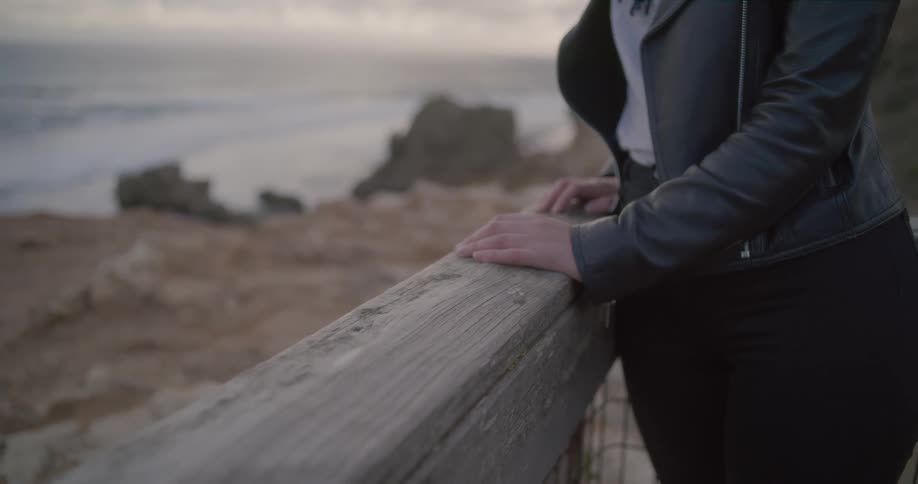 Close-up of a woman's hands on a wooden railing as she contemplates the ocean from a rocky shoreline at dusk