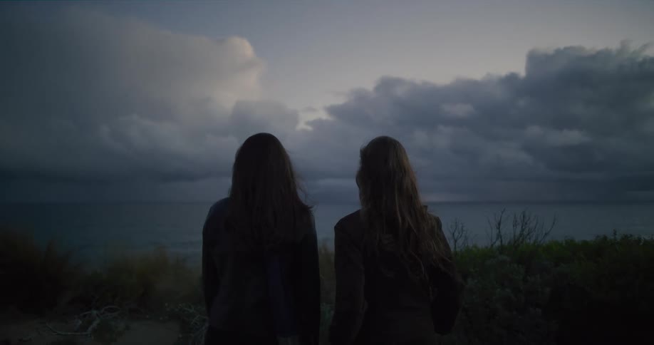 From behind, two women with long hair stand together on a coastal bluff, looking out at the moody ocean and a dramatic, cloudy sky at dusk