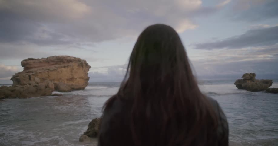 A woman with long dark hair stands on a rocky shore, looking out at the ocean waves during a cloudy evening
