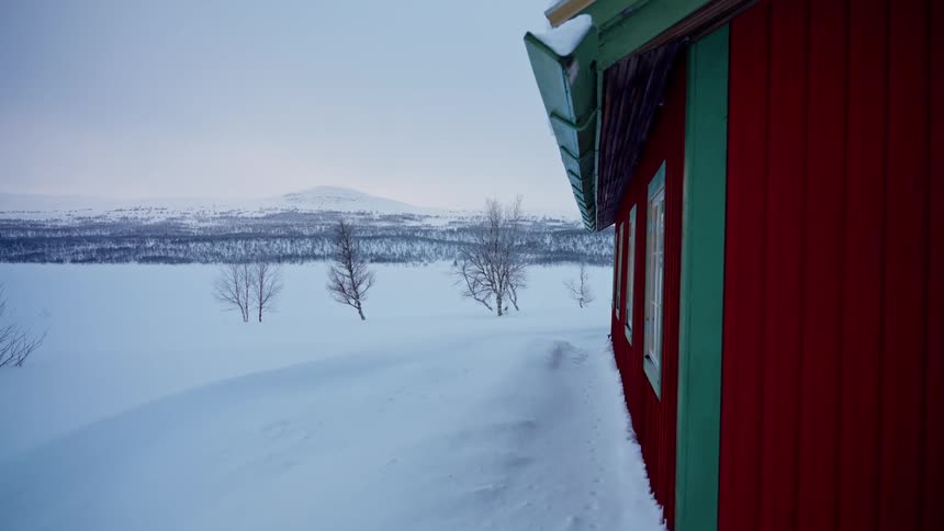 A static shot shows a traditional red wooden house standing in a vast, serene, snow-covered winter landscape