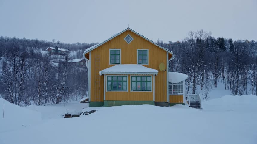 A bright yellow house stands alone in a serene, snow-covered landscape with warm lights glowing from its windows