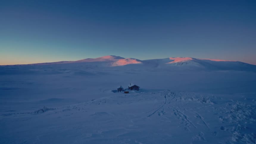 An isolated cabin sits alone in a vast, snow-covered landscape as the sun's first light creates an alpenglow on distant mountains