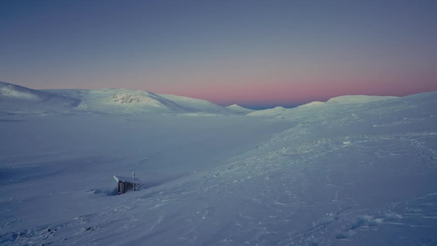 Pastel sunrise colors paint the sky over a vast, snow-covered arctic valley with a solitary wooden hut buried in the snow