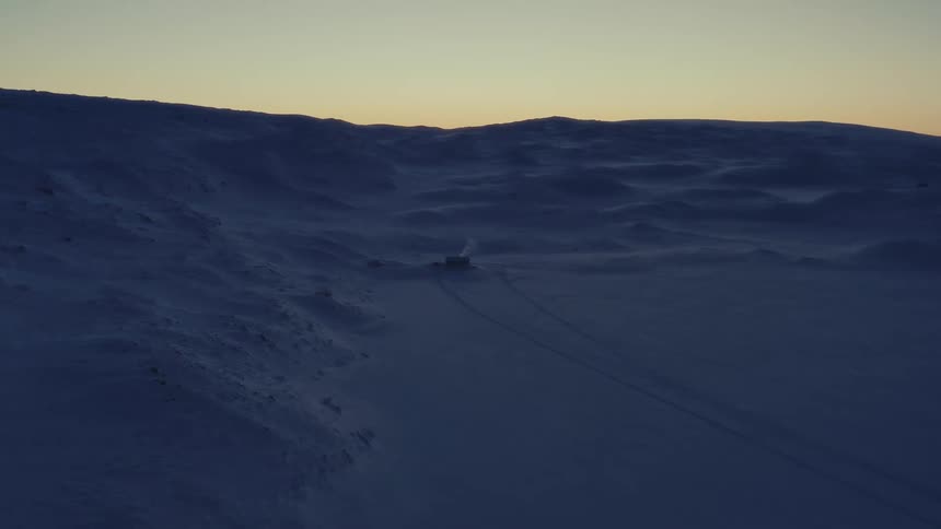 A solitary cabin sits in a vast, desolate, snow-covered landscape at dusk, with smoke gently rising from its chimney