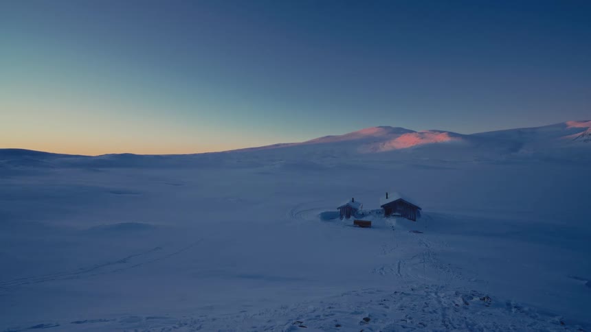 A remote wooden cabin sits isolated in a vast, snow-covered arctic landscape as the last light of sunset glows on the distant mountains