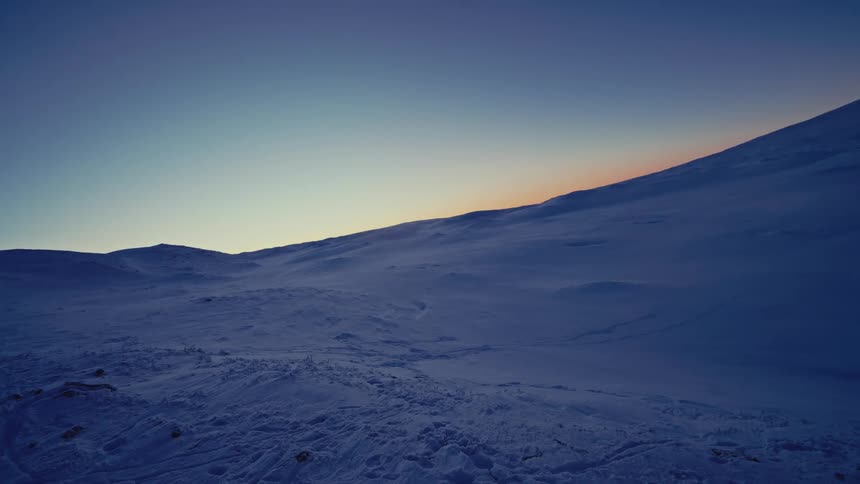 An expansive, snow-covered mountain landscape is slowly revealed under a clear twilight sky