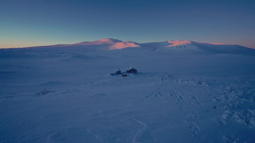 A remote cabin sits alone in a vast, snow-covered landscape while the distant mountains are bathed in the pink glow of sunrise