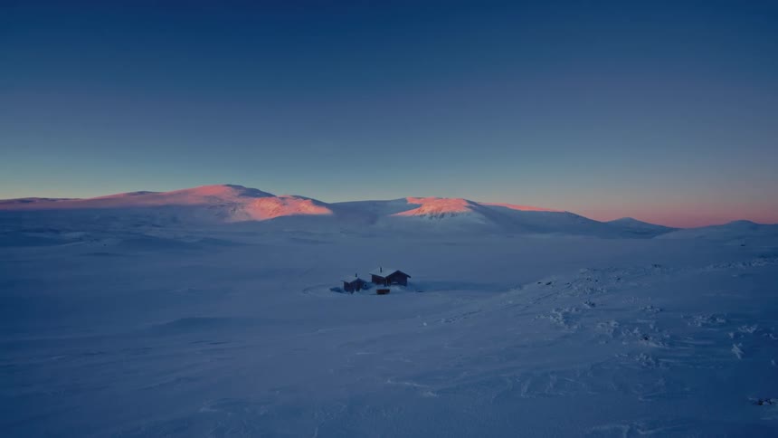 An isolated cabin sits in a vast, snow-covered landscape as the sun casts a pink glow on the distant mountains at sunrise