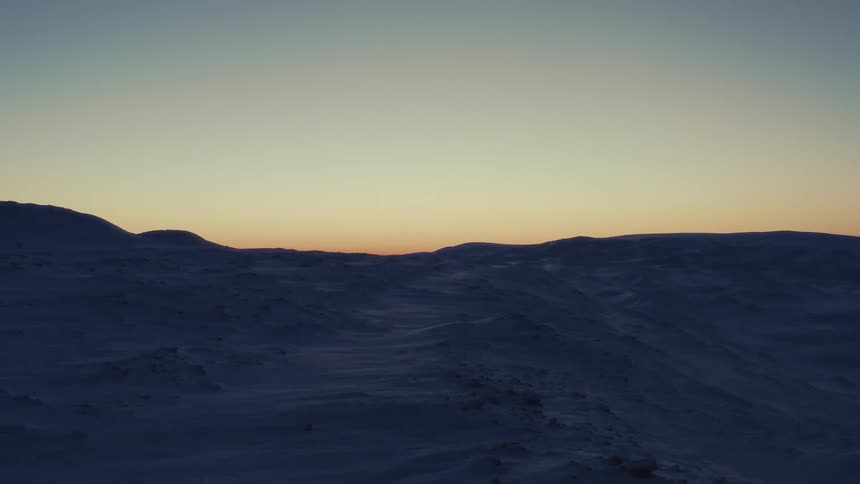 Golden light from a sunrise illuminates the horizon over a vast and windy snow-covered landscape