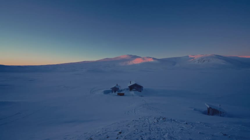 Remote wooden cabins sit peacefully in a vast snowy wilderness as the setting sun casts a pink glow on the distant mountains