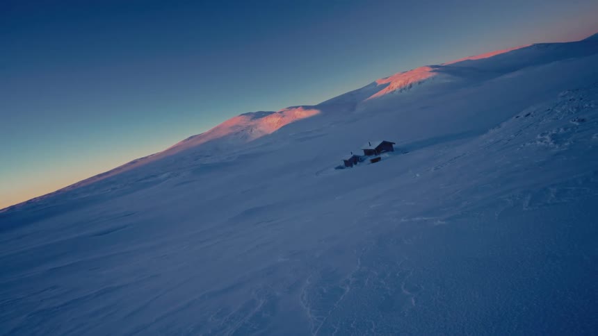 An aerial view reveals remote cabins nestled in a vast, snow-covered mountain landscape as the sunrise casts a warm glow on the distant peaks