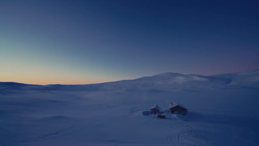 An aerial view of a remote wooden cabin in a vast snowy landscape with smoke rising from the chimney during the serene blue hour
