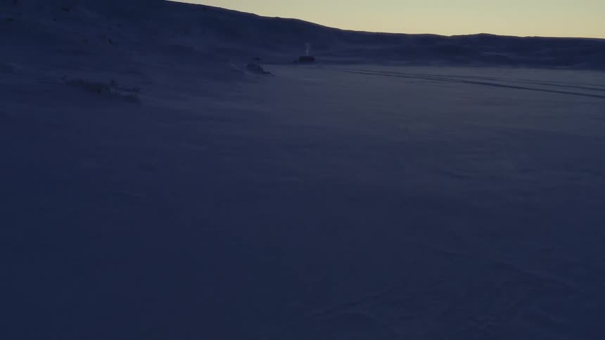 A slow tilt-up shot reveals a solitary cabin with a smoking chimney amidst a vast, desolate, frozen landscape at twilight