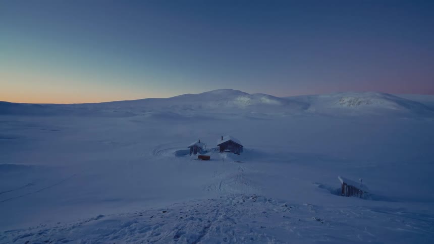 Isolated wooden cabins sit peacefully in a vast, snow-covered mountain landscape at twilight