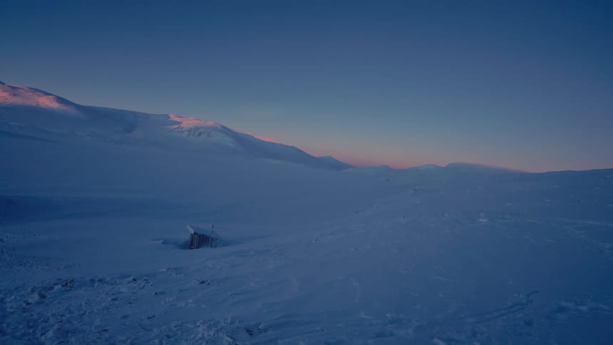 A remote outhouse is half-buried in a vast snowy landscape as the sun sets over the mountains of Sweden