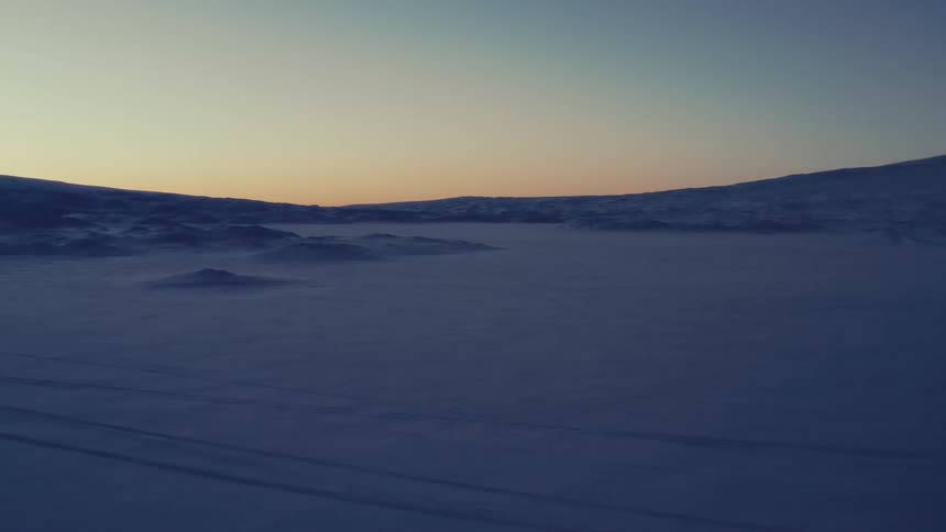 A cold wind blows snow across a vast, frozen arctic landscape at sunrise or sunset