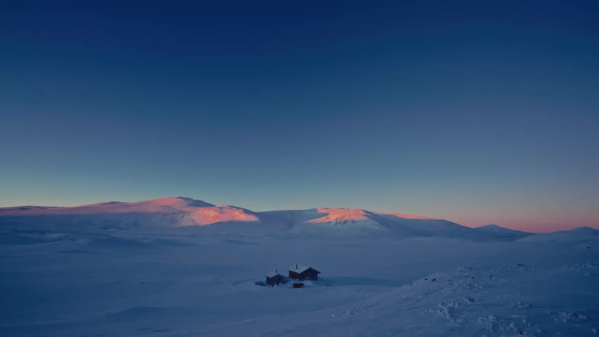 An isolated cabin rests in a vast, snow-covered landscape at twilight as the last rays of sunset illuminate the distant mountain peaks
