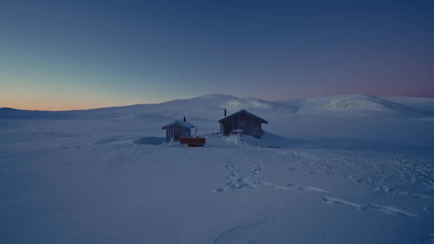 Two remote wooden cabins sit in a vast, snow-covered landscape under a peaceful twilight sky