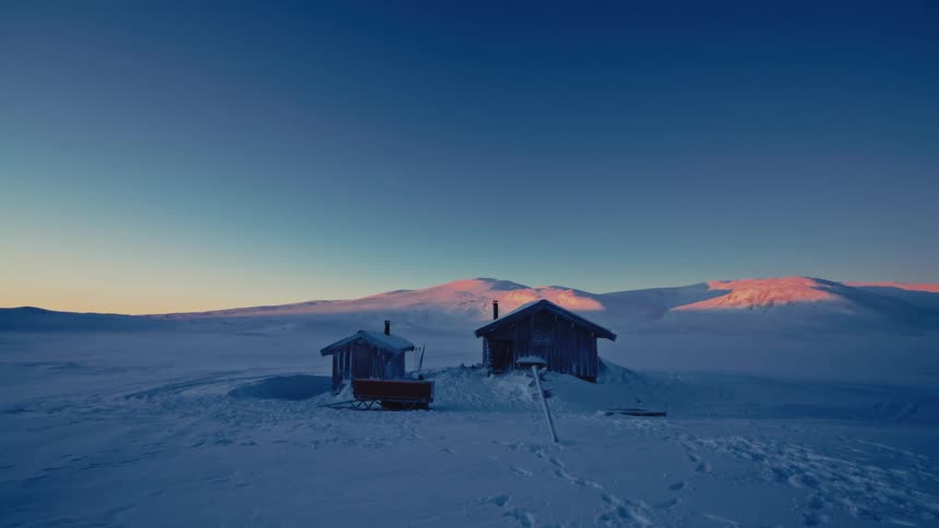 Two remote cabins sit in a vast, snow-covered arctic landscape as the sunrise casts a warm glow on the distant mountains