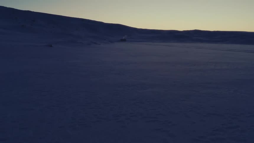 A slow, wide reveal of a desolate, snow-covered arctic landscape at dusk, with a lone cabin in the distance