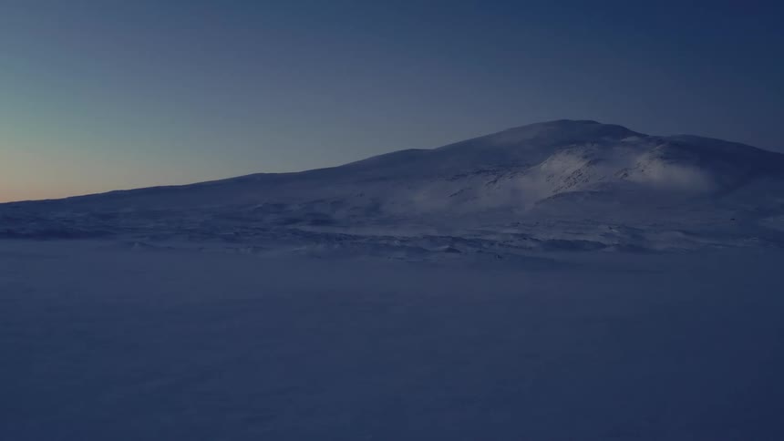 A vast and desolate snow-covered landscape with a mountain silhouetted against a deep blue twilight sky