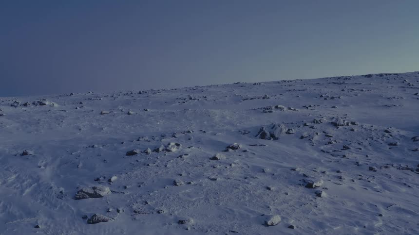 An aerial view reveals a vast, desolate, and rocky tundra landscape covered in snow at twilight