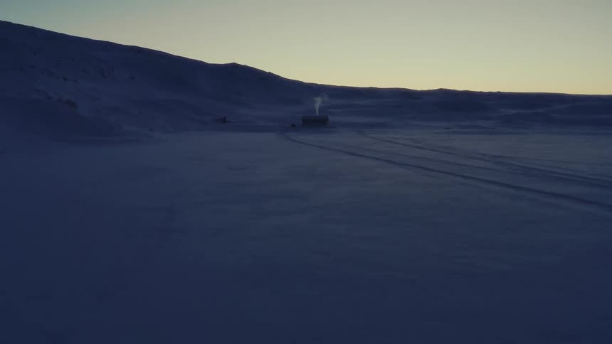Aerial view of a remote cabin nestled in a vast, snow-covered landscape at dusk, with smoke gently rising from its chimney