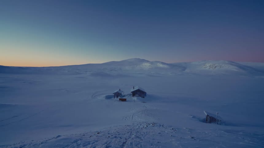 Isolated wooden cabins sit in a vast, snow-covered mountain landscape during a serene winter twilight