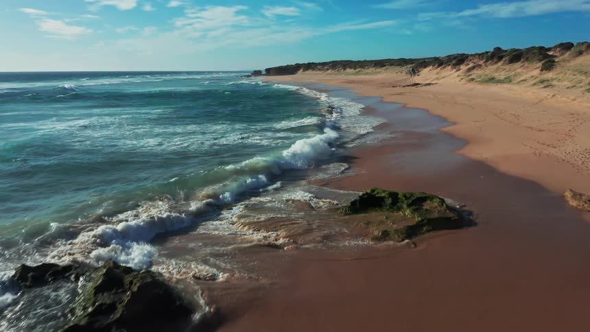 A slow-motion aerial dolly shot glides over the ocean waves as they break along a picturesque and secluded sandy beach