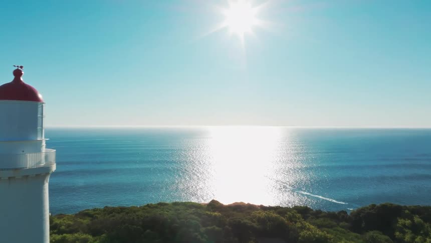An aerial drone shot flies towards a picturesque white lighthouse on a green coastal headland, with the bright sun reflecting on the calm blue ocean