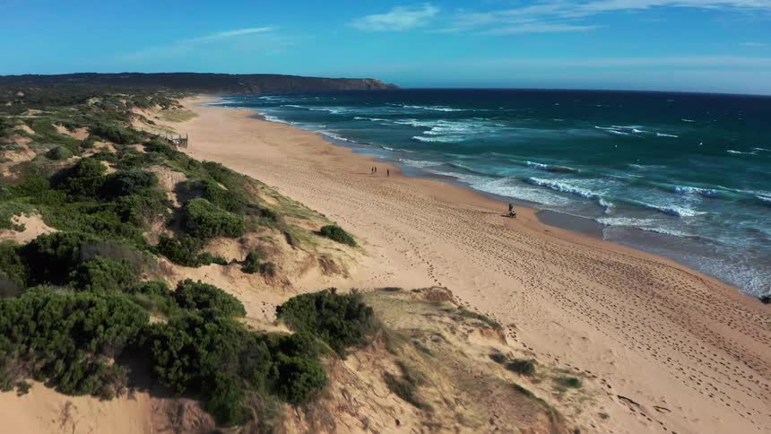 A stunning aerial view flies over coastal sand dunes towards a beautiful sandy beach with people walking along the shoreline