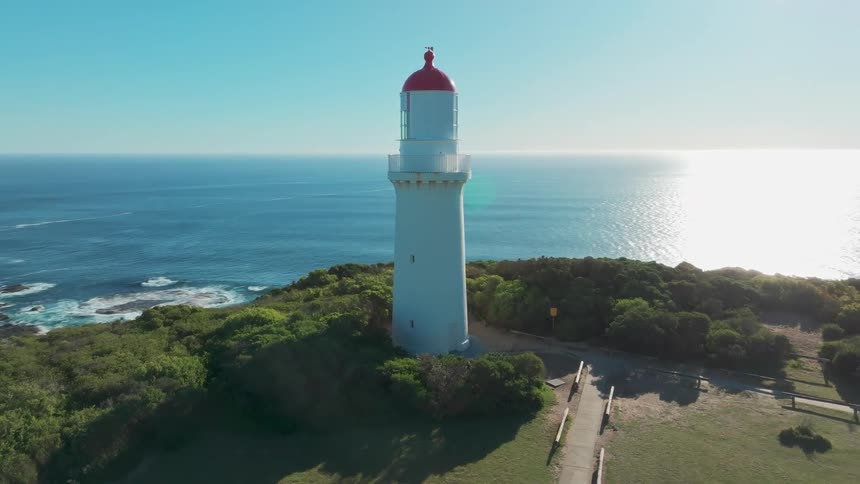 Aerial footage circles a picturesque white and red lighthouse on a lush green cliff overlooking the sparkling ocean on a bright sunny day