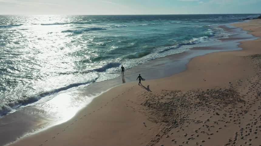 An aerial slow-motion shot follows two surfers with their boards as they walk along a sandy beach next to the shimmering ocean waves on a sunny day