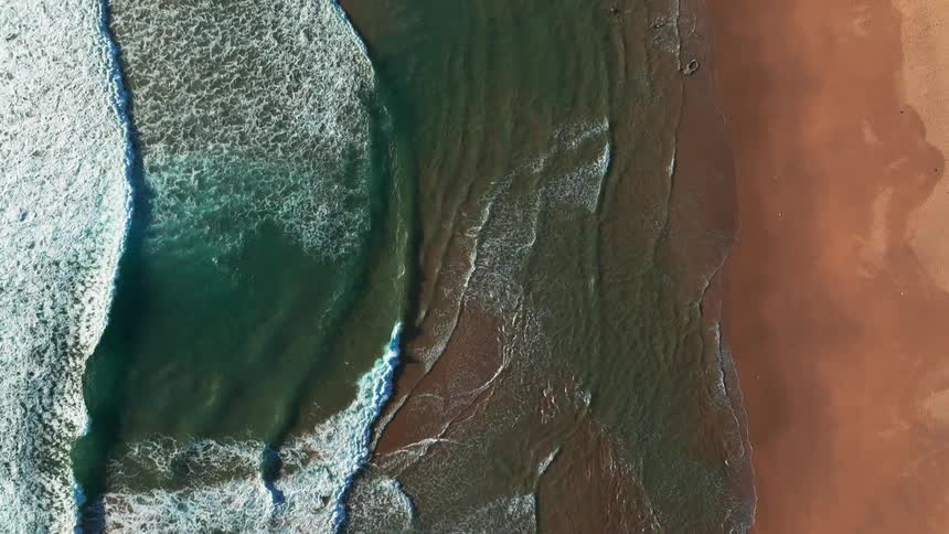 A stunning slow-motion aerial top-down view of ocean waves gently breaking on a sandy beach, revealing submerged rocks as the water recedes