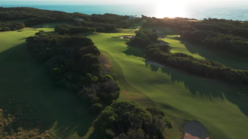 An aerial view flying over a beautiful, lush green golf course with sand bunkers next to the ocean at sunset