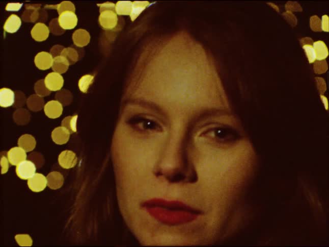 Retro footage shows a close-up of a young woman's face as she smiles then looks seriously into the camera with beautiful bokeh lights behind her