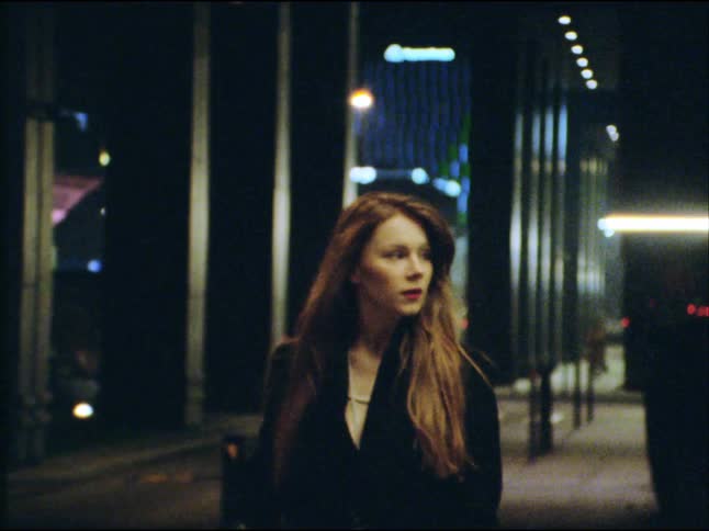 A moody cinematic shot of a young woman with long hair walking down a city street at night