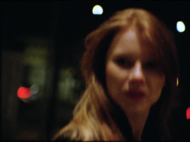 A pensive young woman walks down a city street at night in this moody, handheld shot with out-of-focus bokeh lights in the background